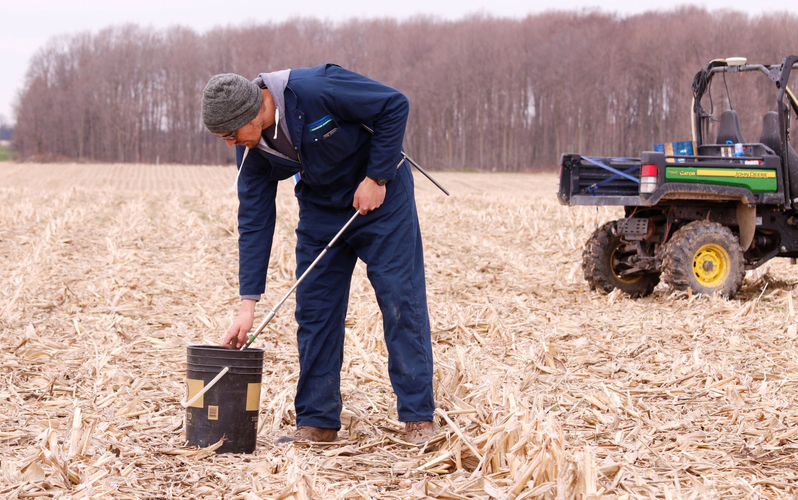 Person standing in corn stubble field, putting a soil sample into a bucket,near an all terrain vehicle