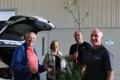 Two customers and two staff pose with a tree behind a van with an open trunk