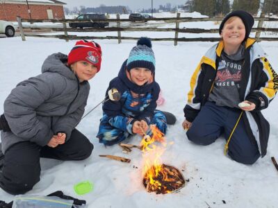 Three kids sit around a small fire in the snow roasting marshmallows