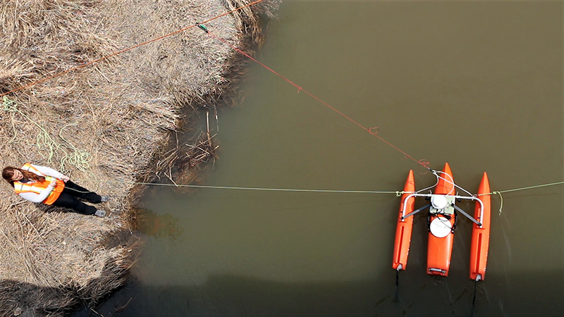 A person stands next to a stream, holding a rope attached to equipment floating in the water.