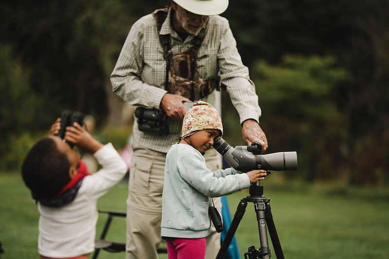 An adult steadies a telescope as a young child looks through it, while a second child looks skyward through binoculars