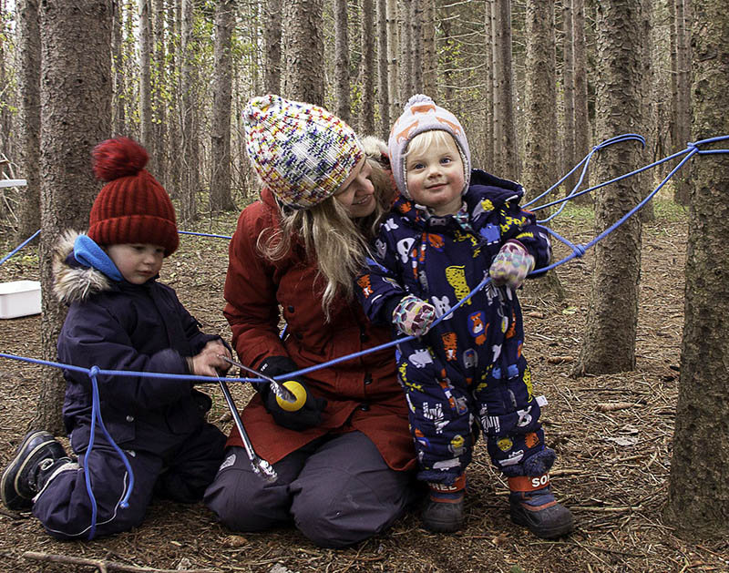 Two children and their caregiver play in the woods at Fanshawe Nature School