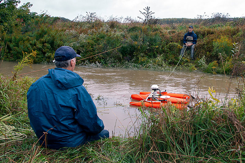 Two staff sit on opposite sides of a creek and guide equipment to measure stream flow back and forth across the creek