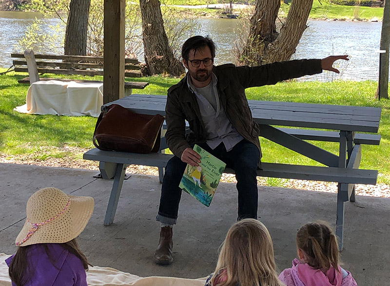 Person sitting on picnic table bench, holding a children's book and talking to children seated on the ground in front of him, with a river in the background