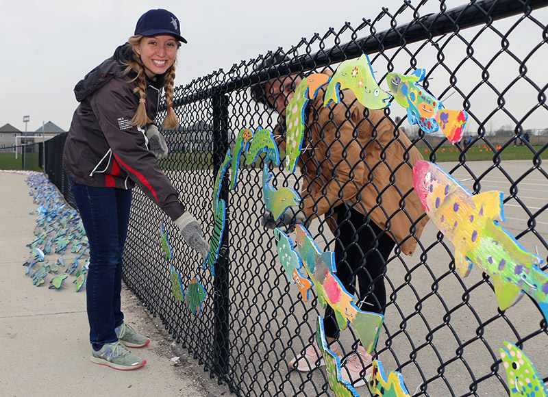 Two people stand next to a chainlink fence where they are mounting painted wooden fish