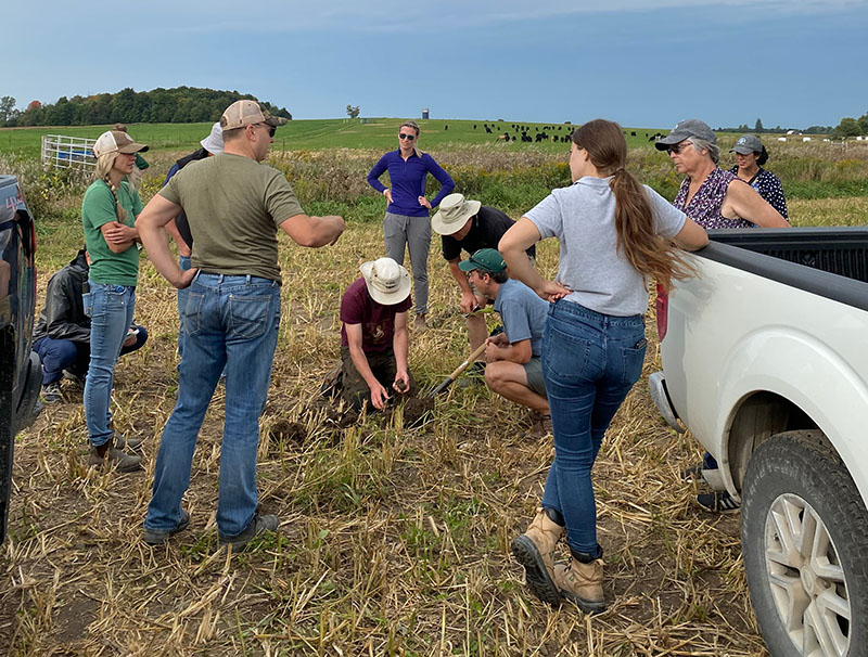Group of people stand in a farm field, looking at people crouching down examing the soil.