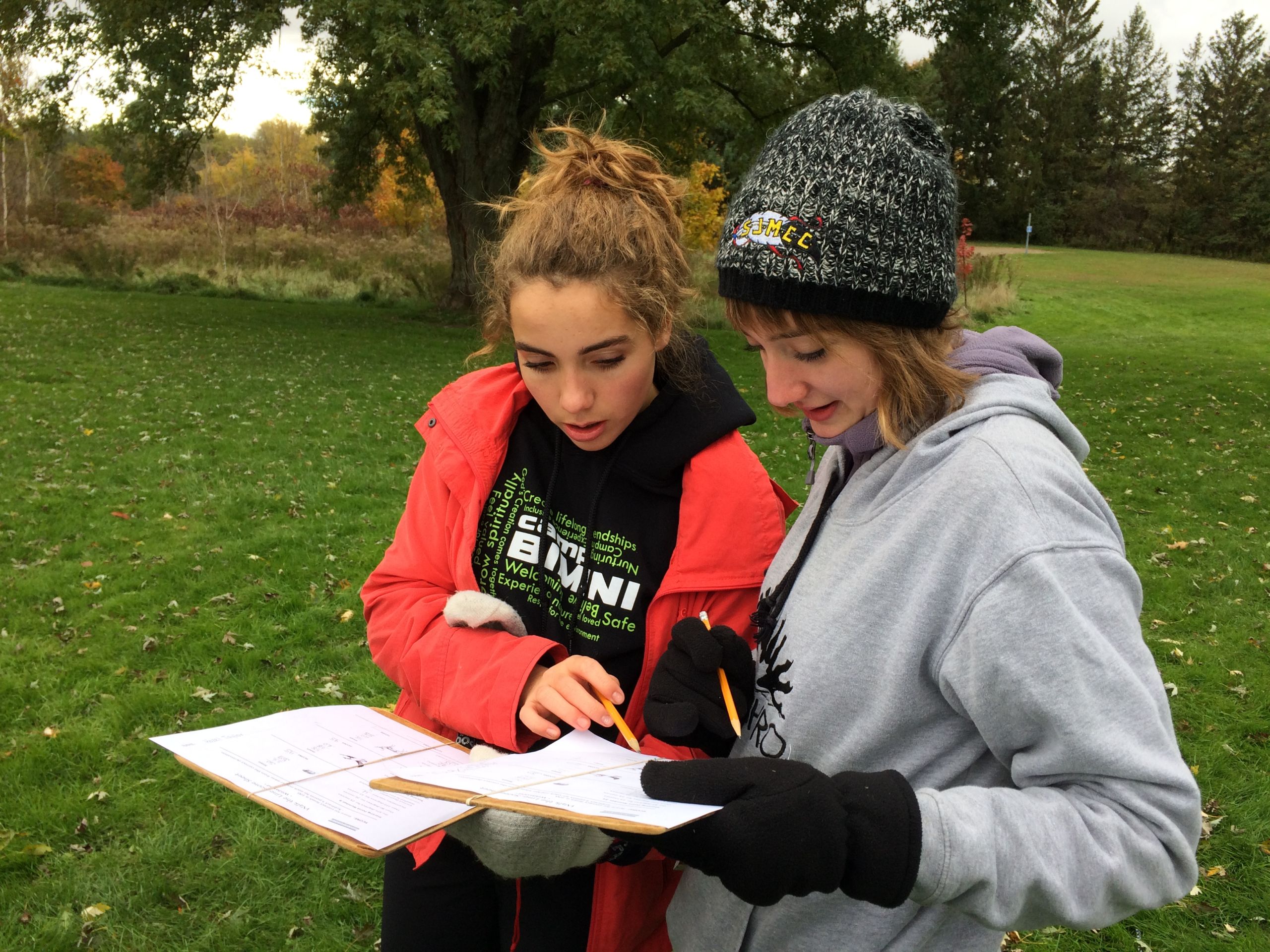 Two students standing in a grassy field, looking at worksheets on clipboards they are holding