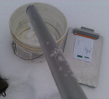 Metal tube lays across a bucket in the snow, next to a clipboard