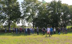 A group of people stand in an agricultural field listening to a presenter