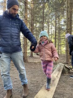 A caregiver helps a young child walk along a balance beam in the forest