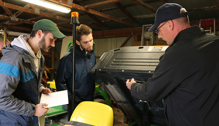 Three people in a mechanics workshop, looking at machinery and making notes on a clipboard