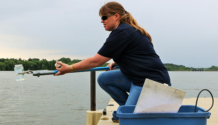 Person kneeling on a wooden dock next to a lake, holding water quality testing equipment