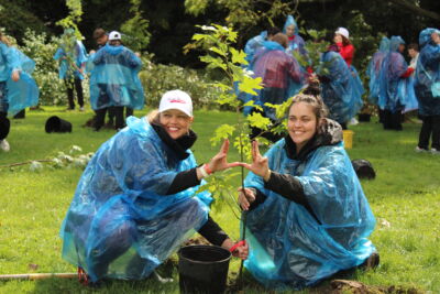 Two female students form a W with their hands over a tree they planted