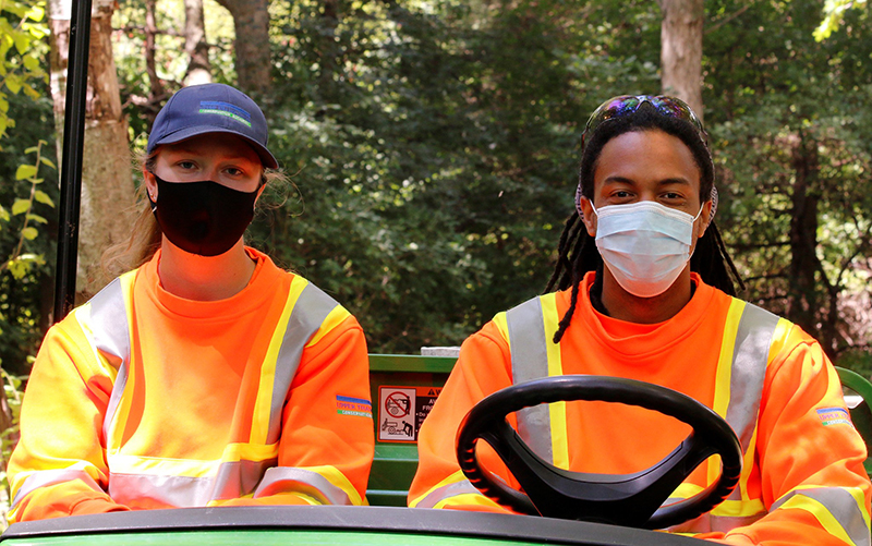 Two people wearing orange and yellow safety uniforms and masks, sitting in a golf cart