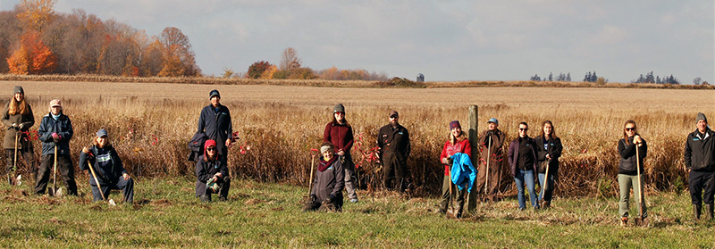 People holding shovels standing in a field with tall grasses and trees in the background
