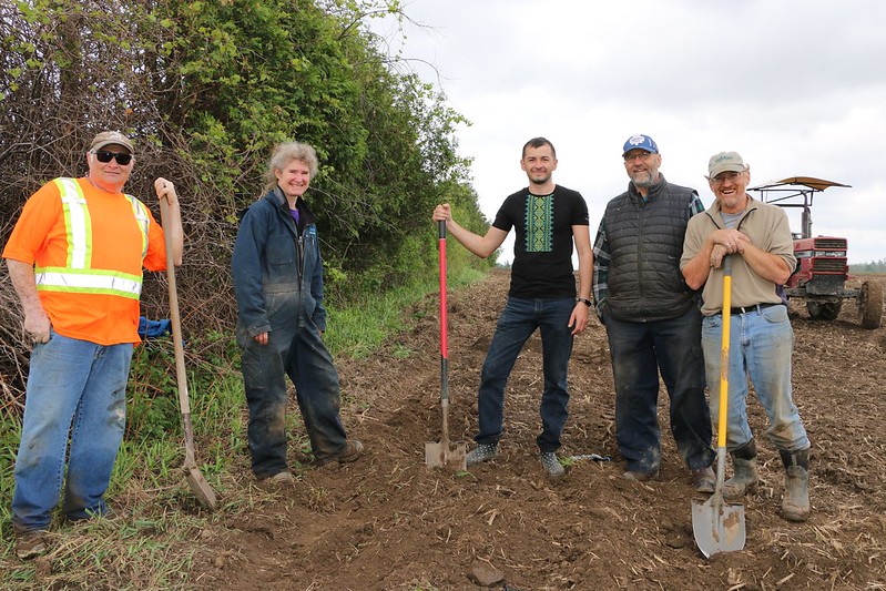 Five people stand with shovels on a dirt field.